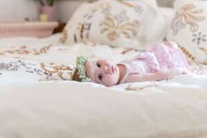 A baby girl wearing a floral crown and pink christening dress lying on patterned bedlinen.