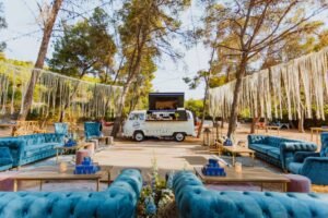 A vintage white mobile bar van parked in a pine forest, featuring blue velvet sofas and wooden coffee tables arranged for a bohemian-style wedding.