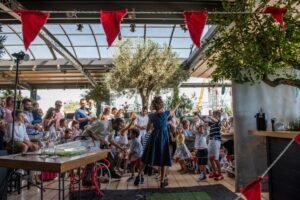 A man wearing an apron and a woman in a blue polka-dot dress perform a La Petite Marguerite bubble show for children at a Gazarte christening reception, with red bunting decorating the outdoor backdrop.