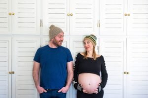 A pregnant woman and a man in beanies look at each other in front of white panelled cupboards during a pregnancy photoshoot in Tinos, Greece.